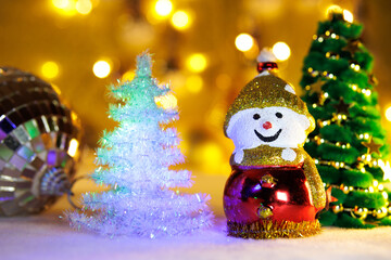 Two Christmas trees with a snowman and a Christmas ball on a blurred background with yellow lights