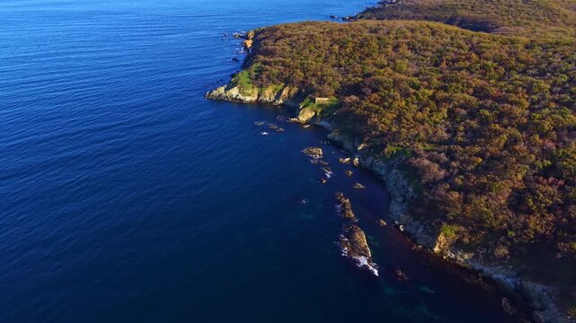 Morning light graces the rugged coastline, illuminating lush greenery and the deep blue ocean. Waves gently lap against the rocks, creating a peaceful atmosphere.