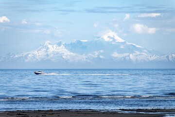 View over Cook Inlet to white mountain peaks of Mount Iliamna, motor boat rides on the ocean, mountains of the Aleutian Range, Anchor Point, Anchor River State Recreation Area, Alaska, USA