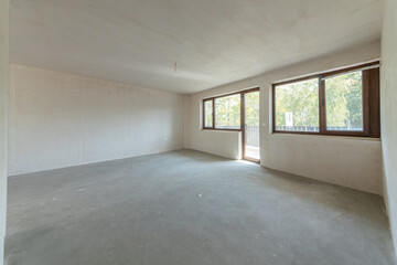 Interior of an empty room under construction in a new apartment. Unfinished space with raw concrete walls, floor, and a large window. Renovation and real estate concept.
