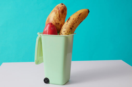 Miniature trash bin with spoiled fruits and vegetables on a blue background