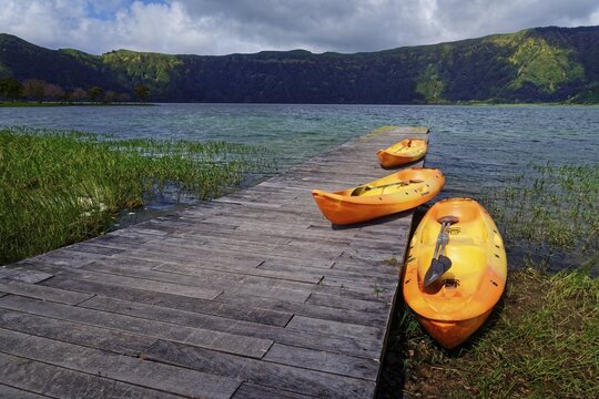 Three orange kayaks on the wooden jetty of the crater lake Lagoa Azul, surrounded by greenery and under a slightly cloudy sky, crater circular hiking trail, Caldeira das Sete Cidades, Sete Cidades, Lagoa Azul, Sao Miguel, Portugal