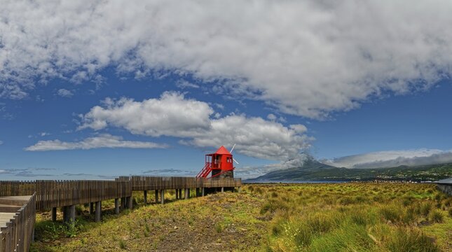 Wooden path leads to the Moinho das Lajes windmill with a dramatic sky and the volcanic cone of Pico in the background, fishing village, Lajes do Pico, Lajes, Pico Island, Azores, Portugal