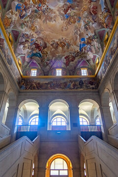 The grand Great Staircase of El Escorial in Spain features massive granite arches leading to a stunning vaulted ceiling covered in vibrant Renaissance frescoes