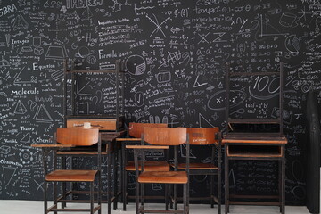 Classroom with empty wooden chairs and desks against a black chalkboard filled with mathematical equations, formulas, and scientific diagrams in a learning environment