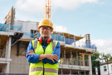 Portrait of woman builder with ear defenders and arms crossed stands confidently at building site, looking over ongoing work as cranes operate in background