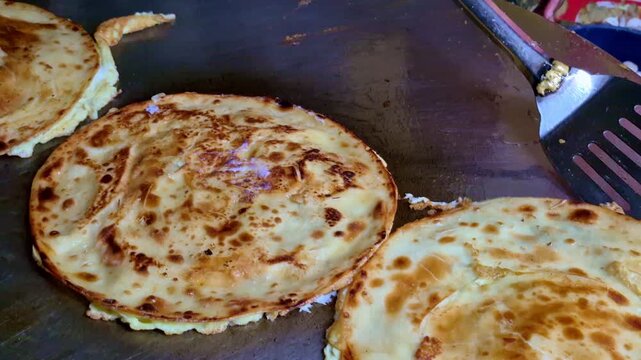 A close-up of multiple eggroll parathas cooking on a large flat griddle, showing evenly browned surfaces, flaky layers, and a metal spatula ready for flipping.