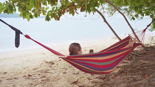 A Woman Relaxing in a Hammock beside a Quiet Sandy Shore while Looking at her Smartphone under Shaded Branches with a Peaceful Coastal Horizon creating a Calm Beach Travel Atmosphere