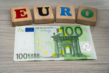 one hundred euro banknote lying on a wooden table below wooden blocks spelling the word euro for financial concepts, banking, and european economy themes in 2026
