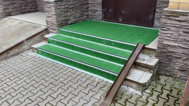 Green Turf Covered Outdoor Steps At Stone Entrance With Metal Handrail, Interlocking Pavers And Worn Edges, Subtle Moss Along Joints, Maintenance Access To Rooftop Service Area, Muted