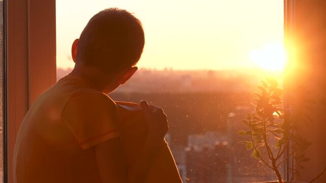 Sad pensive child sitting on windowsill and looking through window at city during beautiful golden sunset. Little boy feeling lonely and dreaming about future in moment of solitude and reflection