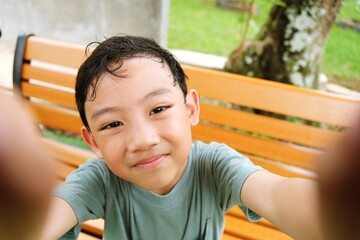 Smiling young boy taking a selfie while sitting on a park bench, surrounded by greenery. Represents childhood joy, nature connection, and sustainable lifestyle.