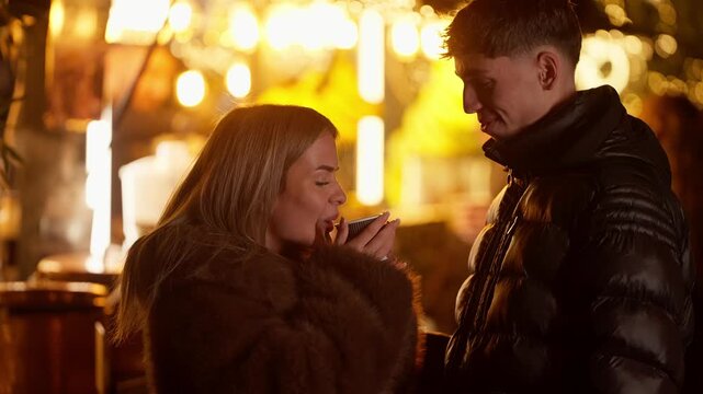 A romantic couple in Paris France are seen drinking a hot beverage at an outdoor Christmas market stall at night The handheld shot captures the warmth and love between the couple amidst the festive li