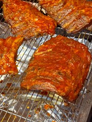Multiple racks of barbecue pork ribs resting on a metal wire cooling rack, likely in a commercial kitchen or deli setting. Vertical view.