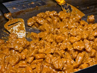 A large quantity of meat chunks being cooked in a thick, creamy orange-brown gravy sauce on a flat-top griddle.