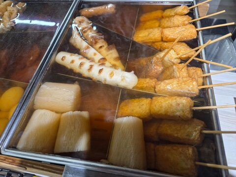 Top-down view of assorted Japanese oden ingredients simmering in a divided stainless steel pot, including daikon radish, grilled fish cakes, and skewered fried tofu in light and dark broths.