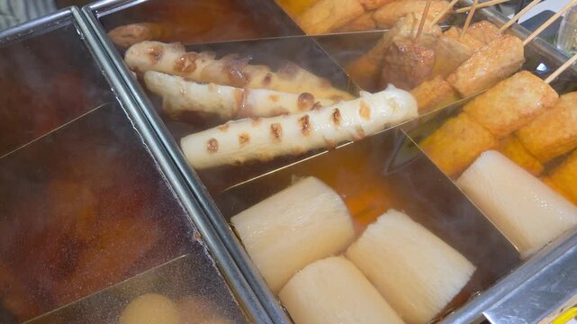 Japanese oden street food display, showing a variety of classic ingredients simmering in a traditional divided metal pot with steam rising.
