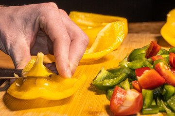 Fresh red, green, and yellow bell peppers are being chopped with a knife on a wooden cutting board. The scene shows healthy cooking, food preparation, and colorful vegetables in a kitchen setting.