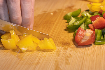 Fresh red, green, and yellow bell peppers are being chopped with a knife on a wooden cutting board. The scene shows healthy cooking, food preparation, and colorful vegetables in a kitchen setting.