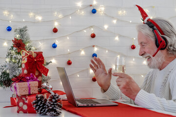 Portrait of old white-haired senior man wearing headphones using laptop to video call with distant people. He celebrates the Christmas event with a glass of sparkling wine and smiling