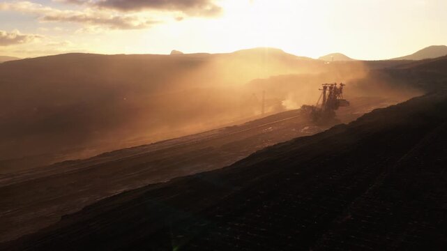 Aerial flyover of an active open-pit coal mine against backlight at sunset, passing massive mining excavators and coal transport machines, highlighting scale, dust, and industrial power.