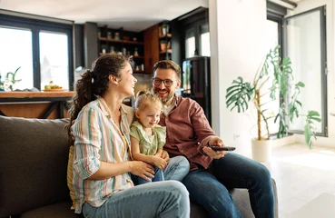 Fotobehang Ridders Portrait of mother, father and daughter having fun holding a remote control and watching tv together at home, family life, parenting, love and bonding concepts  © Lumos sp
