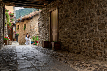 Family is walking down a narrow alleyway with a stone wall on either side village of Potes. The alleyway is dimly lit, and the couple is the only visible figures. Scene is quiet and intimate