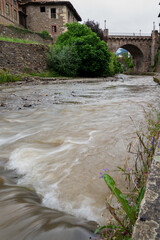 A river is flowing through a town with a bridge over it. The water is murky and the bridge is old