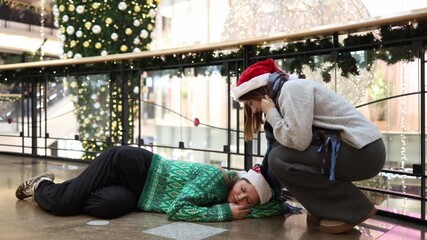 Young woman in santa hat sleeping on office floor after wild new year party while her coworker crouches nearby showing care concern and emotional support