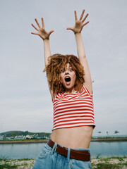 Energetic young woman with curly hair raises arms in excitement, wearing a striped crop top and denim jeans, outdoors by a calm body of water under a bright sky.