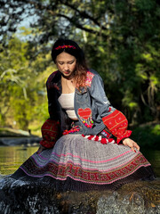 Joyful Asian woman in colorful Hmong fusion dress sitting by a scenic river.
