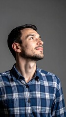 Fototapeta premium Man gazes upward in a blue plaid shirt against a plain grey backdrop, studio lighting illuminates his face