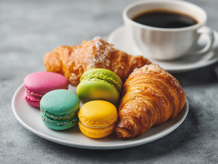 Freshly baked golden croissants paired with colorful assorted macarons served on a white plate accompanied by a steaming cup of black coffee on a grey surface