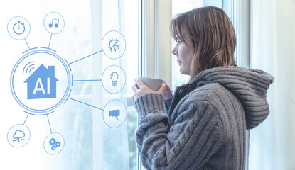 Young woman wearing a grey knit sweater holding a cup of hot drink stands by a window in her smart home controlled by an artificial intelligence system that manages heating and lighting automation