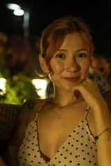 A portrait of a stunning young woman sitting at a cozy outdoor cafe table, drinking coffee during a warm summer evening. Soft ambient lighting, lifestyle photography, and a relaxed atmosphere.