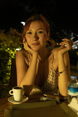 A high-quality lifestyle shot of an attractive young woman sitting at an outdoor cafe during a warm summer evening, enjoying a cup of coffee and smoking a cigarette. 