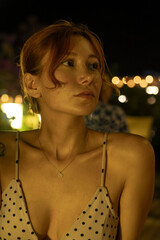 A portrait of a stunning young woman sitting at a cozy outdoor cafe table, drinking coffee during a warm summer evening. Soft ambient lighting, lifestyle photography, and a relaxed atmosphere.