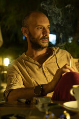 A mature man sitting at a cozy street cafe during a warm summer evening, enjoying a cup of coffee and a cigarette. He is portrayed in a relaxed, urban lifestyle setting with soft ambient lighting