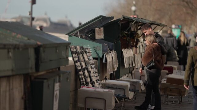A romantic couple is seen browsing books at a traditional book stall along the Seine River in Paris France The scene captures the essence of Parisian life with lovers booksellers and the city's iconic