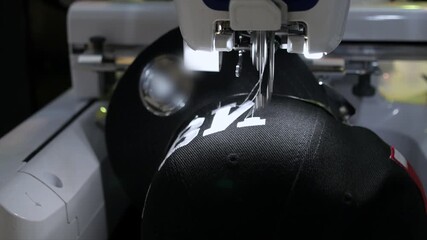 Close up view of a modern industrial sewing machine precisely embroidering a white logo onto a black baseball cap in a textile factory, showcasing automated apparel manufacturing technology