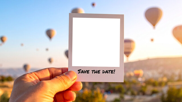 Hand holding an instant photo with 'Save the Date' message. The background shows a stunning sunrise landscape with many hot air balloons flying, creating a romantic and adventurous travel scene. - Powered by Adobe