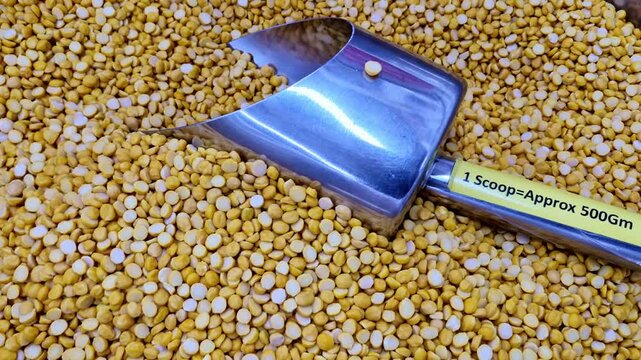 A close-up of split yellow lentils chana daal spread evenly with a metal measuring scoop placed on top, highlighting bulk grains commonly sold in open market displays.