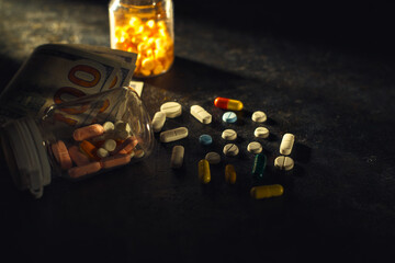 A dramatic, low-key image captures various colored pills and capsules scattered on a dark surface near an overturned prescription bottle and US currency, symbolizing healthcare costs or illicit trade.