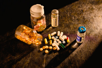 Various colorful pharmaceutical pills and capsules are scattered across a dark surface next to open prescription bottles and labeled vials, including one marked ANTIBIOTICS.