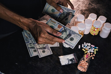 Close-up view of hands handling US hundred dollar bills alongside numerous prescription bottles, loose capsules, and a plastic bag containing white powder.