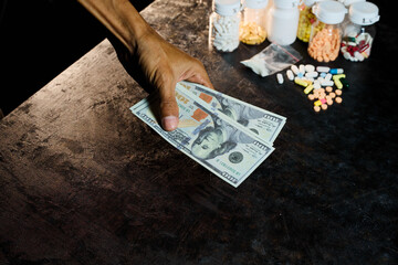 A dark, dramatic image captures a hand offering US hundred-dollar bills near scattered colorful pills and small plastic baggies of white powder, suggesting illicit commerce or substance abuse issues.