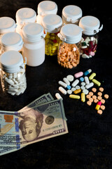 Multiple containers of various pills and scattered colorful tablets lie next to several hundred dollar bills, representing the high cost of pharmaceuticals.