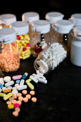 Numerous colorful tablets and capsules are scattered across a dark surface, originating from a tipped-over clear bottle, with several other filled prescription bottles visible in the background.
