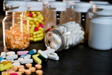 A close-up of assorted colorful pharmaceutical pills and capsules scattered across a dark surface, flowing from an overturned clear glass bottle, sealed prescription containers in the background.