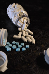 White oblong prescription medication tumbles from a clear plastic bottle, mixing with scattered blue round tablets on a dark, textured surface, symbolizing pharmaceutical treatment and healthcare.